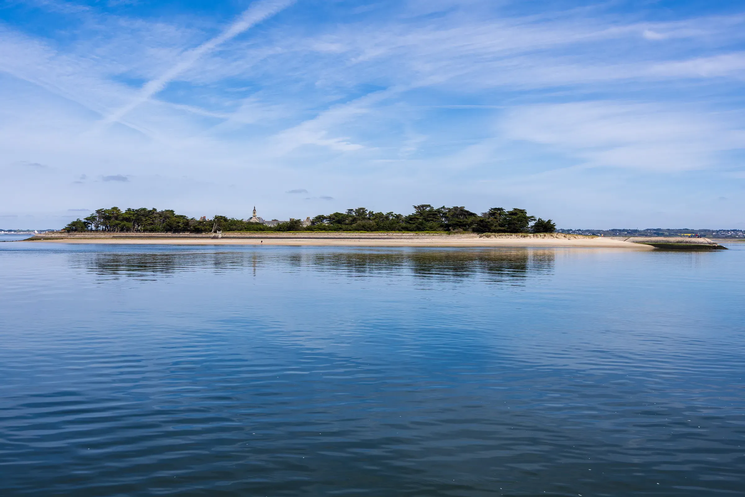 A low flat island covered in trees with a small chapel visible, reflected in calm blue water, Le Croisic