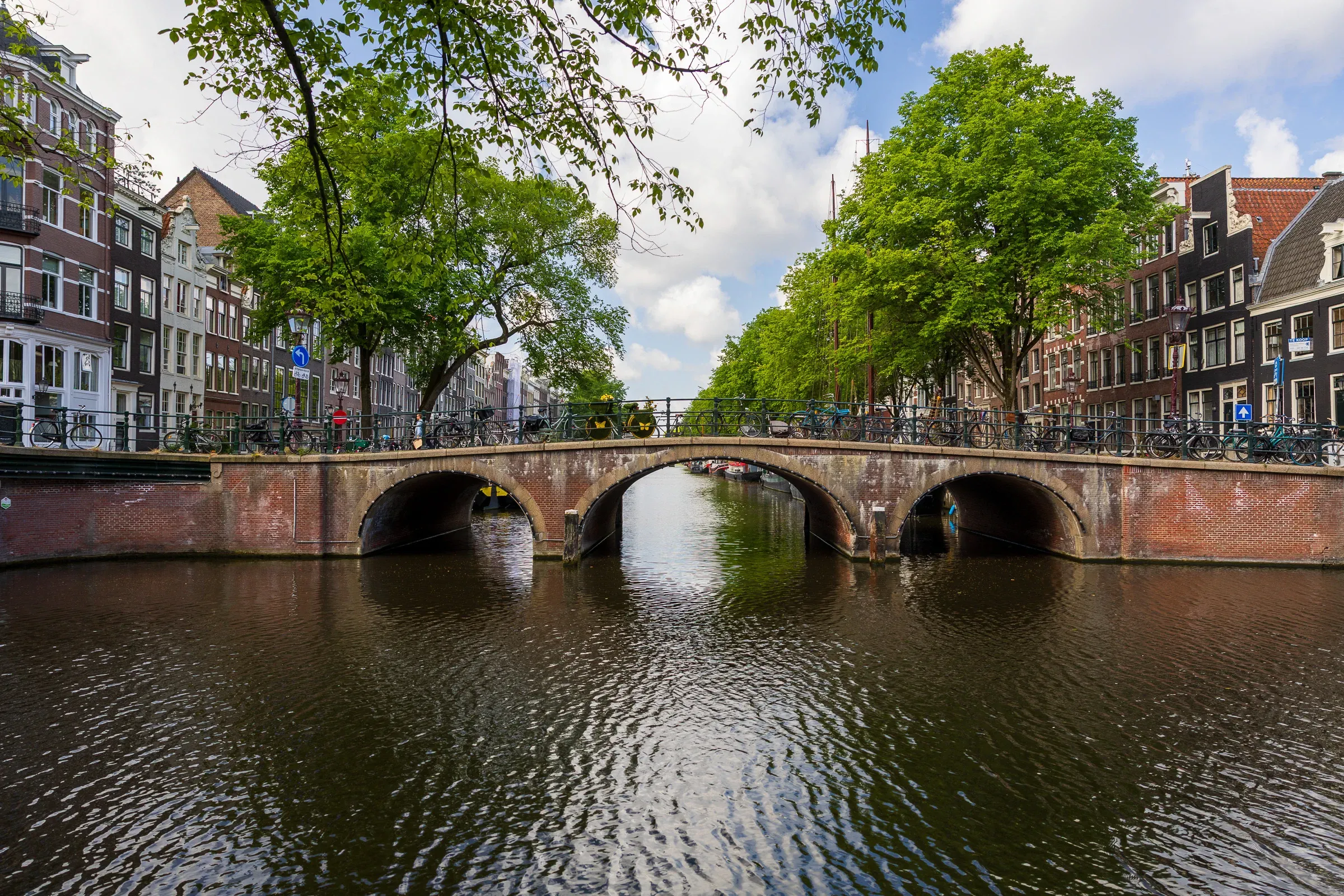 Double-arched brick canal bridge lined with green trees, Amsterdam canal houses reflected in still water