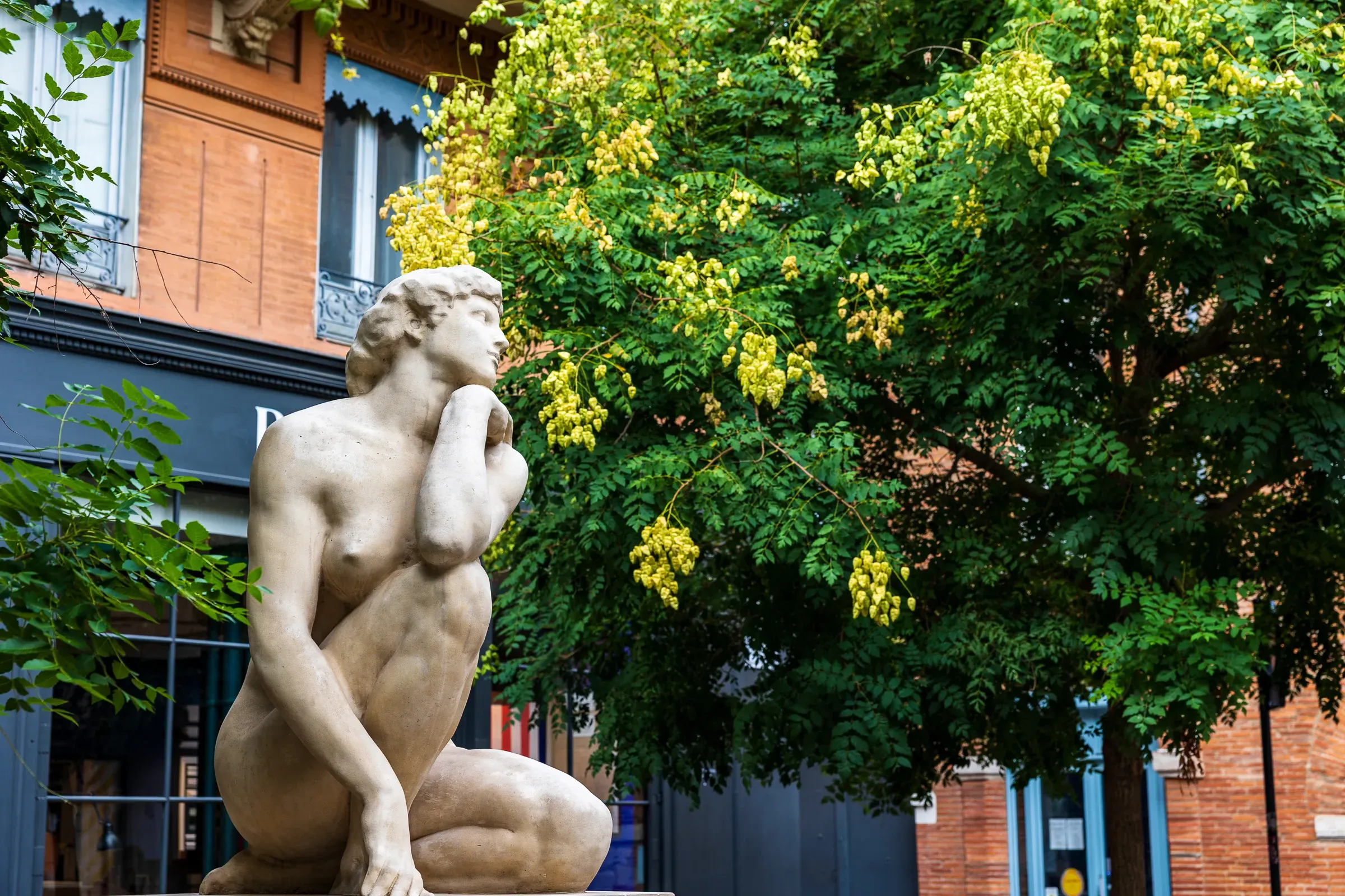 A classical stone sculpture of a crouching figure resting its cheek on one hand, set against a blooming yellow laburnum tree and a pink brick building, Toulouse