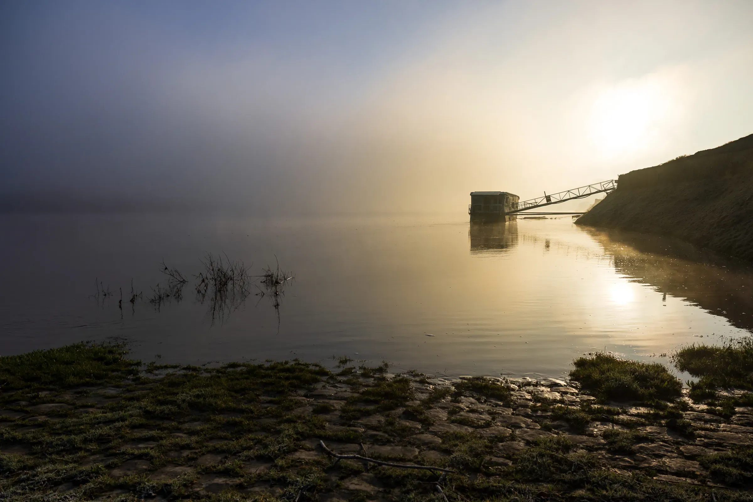 Misty Loire river at dawn, a wooden fishing hut on stilts silhouetted against a sun halo, cobblestone bank with grass patches in the foreground, Saumur