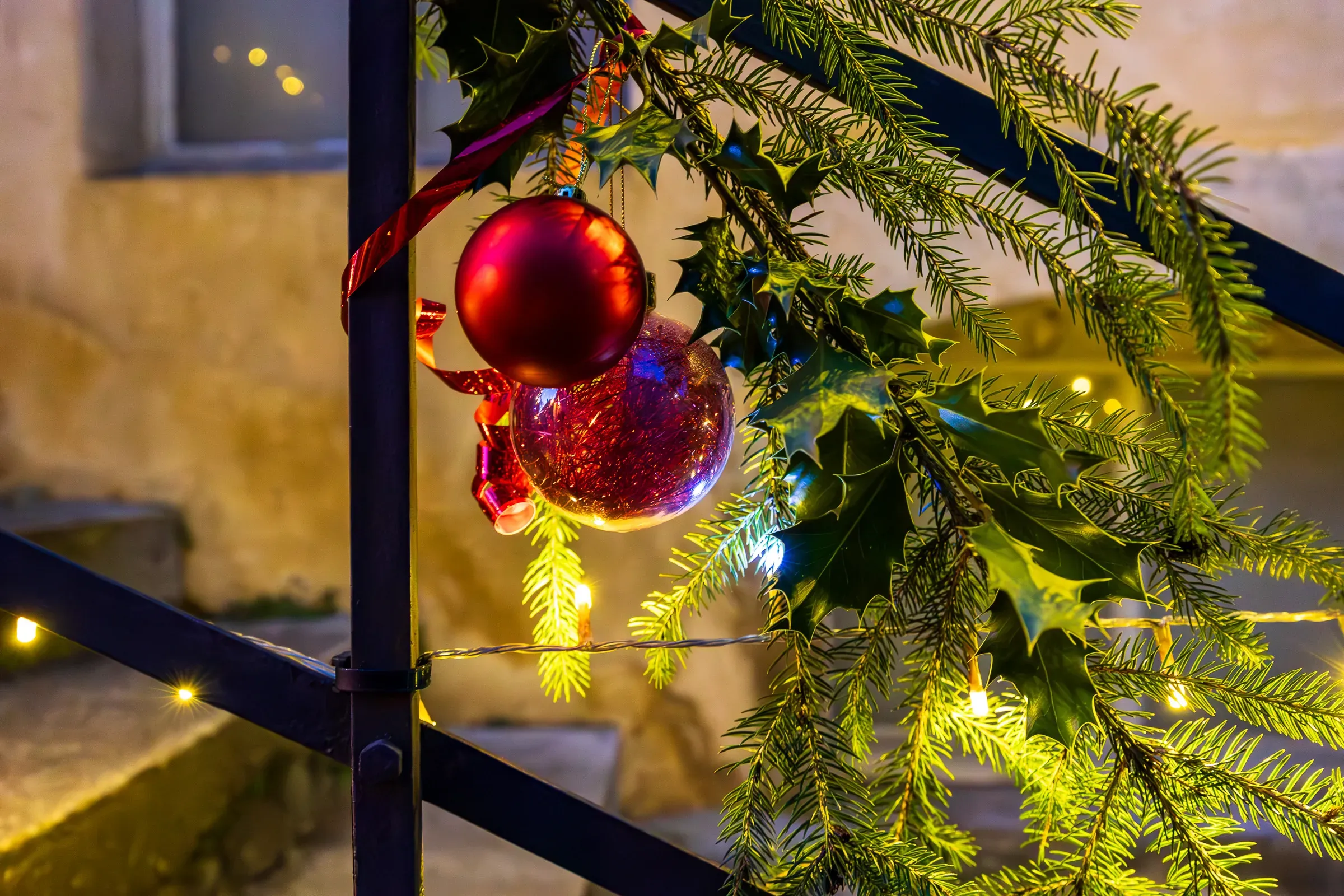 Red Christmas ornaments hanging from fir and holly branches lit by fairy lights, warm amber stone steps behind