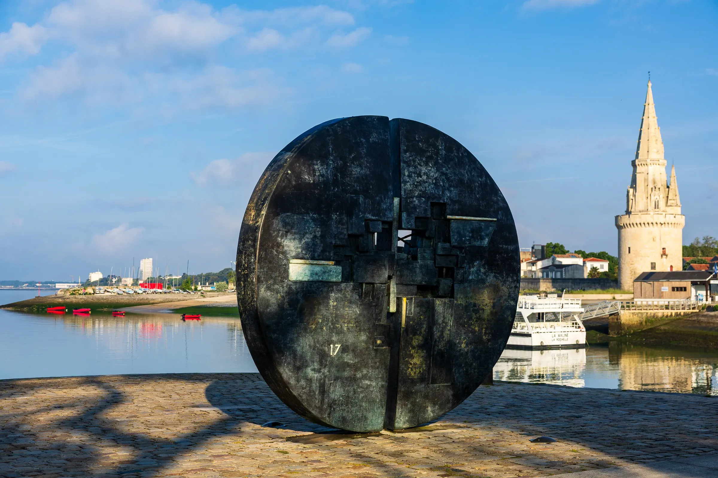 Large circular bronze disc sculpture on the La Rochelle waterfront, the Lanterne tower rising behind it, sailboats and blue sky