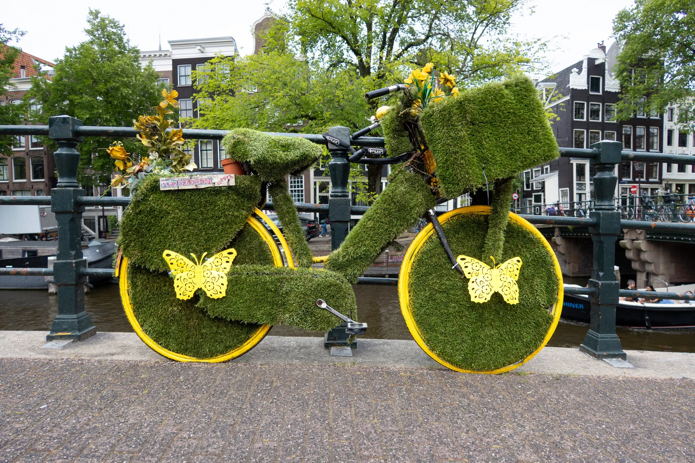 A bicycle entirely covered in green moss with yellow butterfly motifs and yellow wheels, parked on an Amsterdam canal bridge