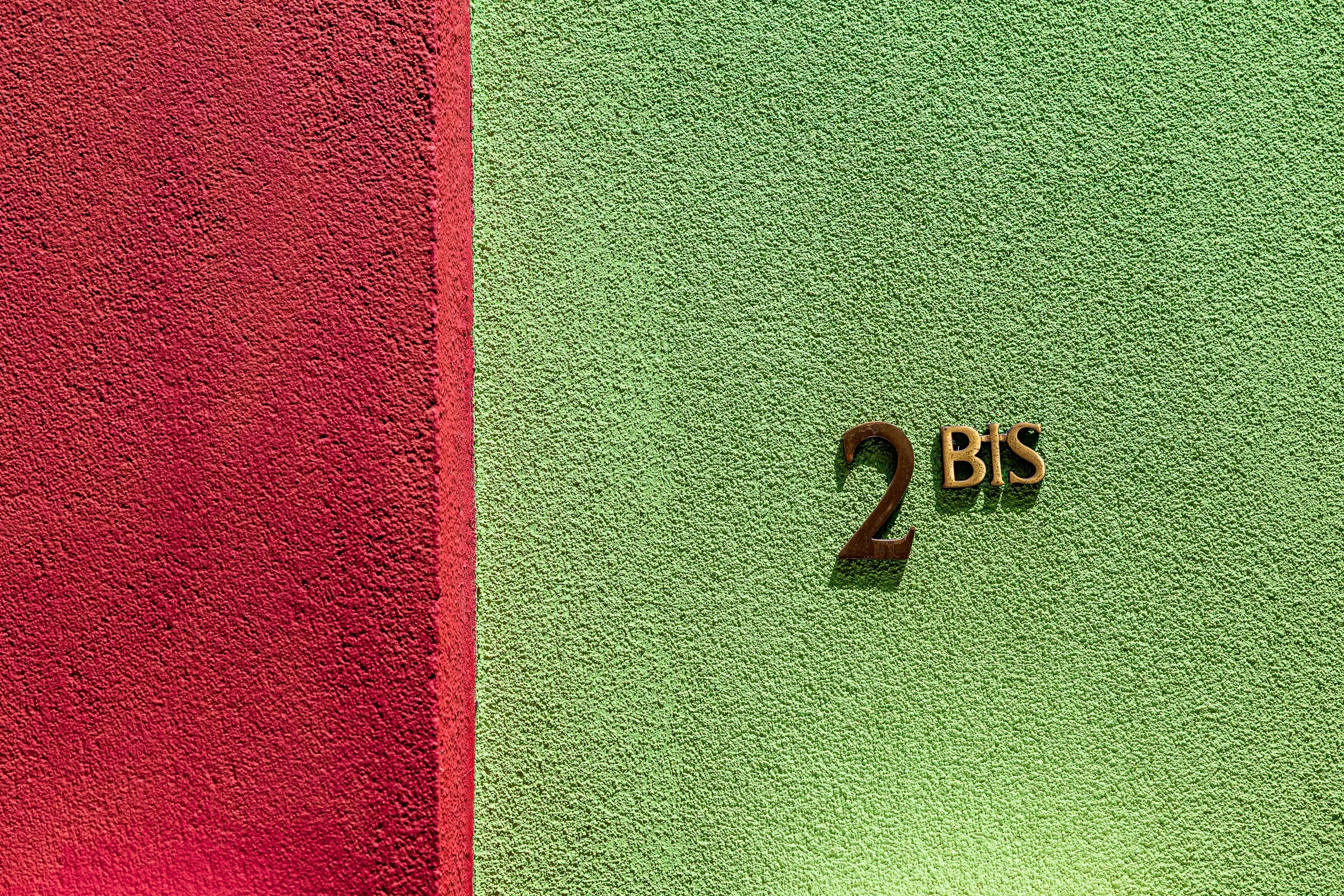 Two adjacent rendered walls — deep red on the left, lime green on the right — with a brass 2 BIS house number, Trentemoult