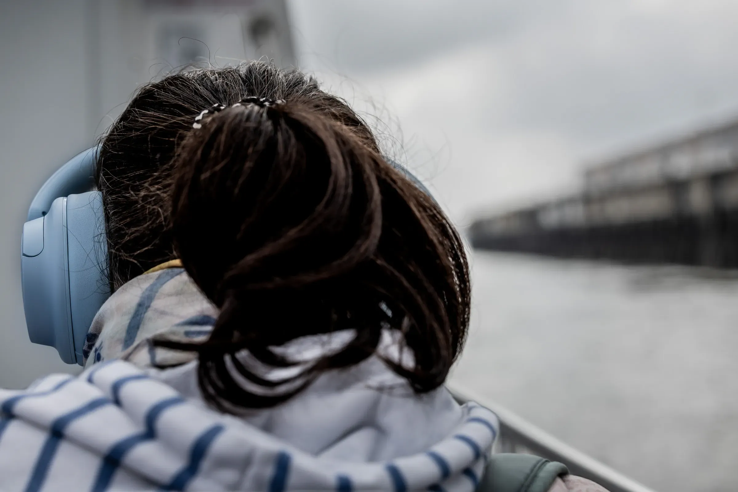 Woman seen from behind with dark hair and light blue headphones, seated outdoors by the Loire, overcast sky
