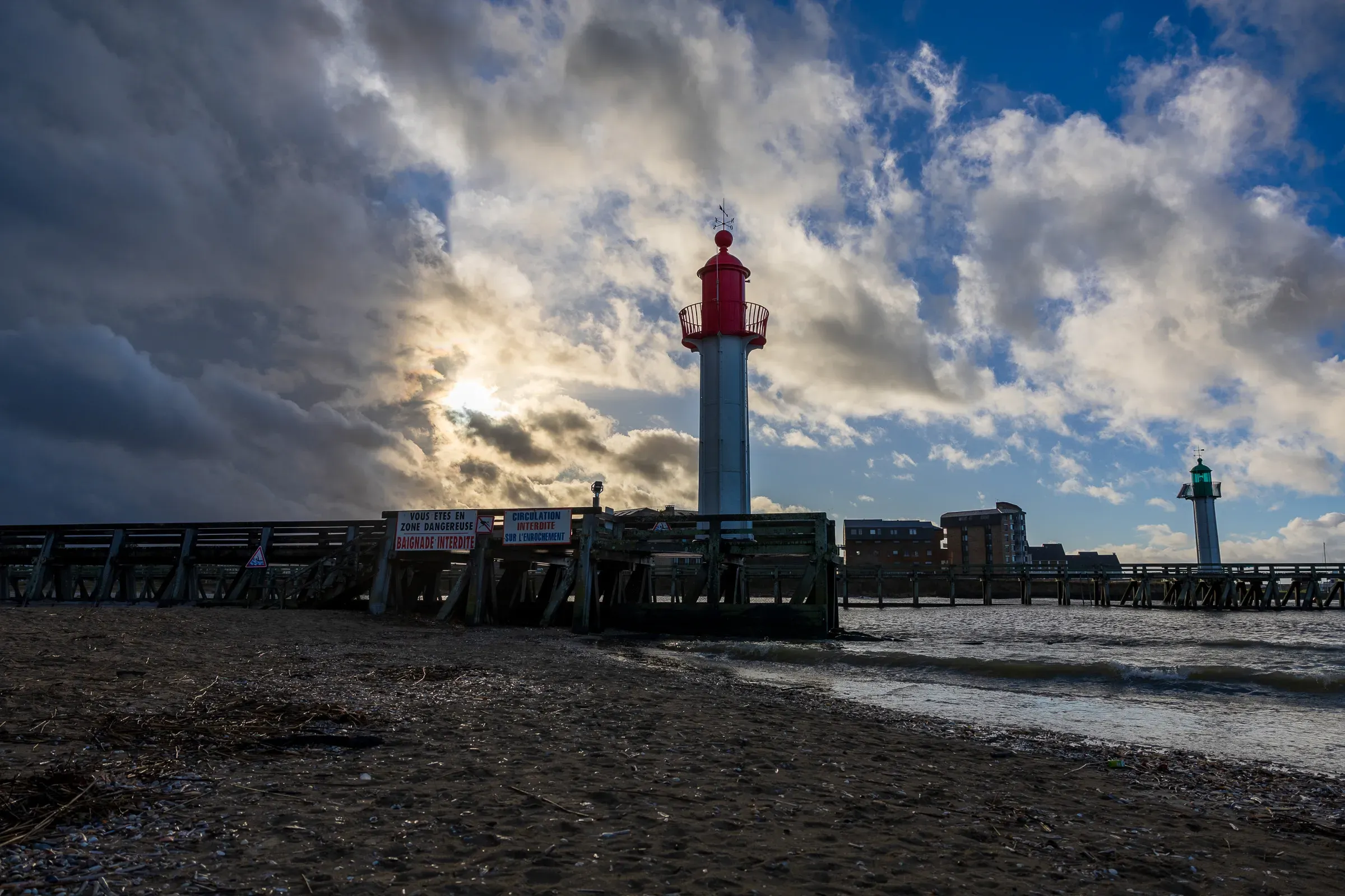 Red and white lighthouse at the end of Trouville's pier, sun breaking through storm clouds, wet sand beach in the foreground, Trouville