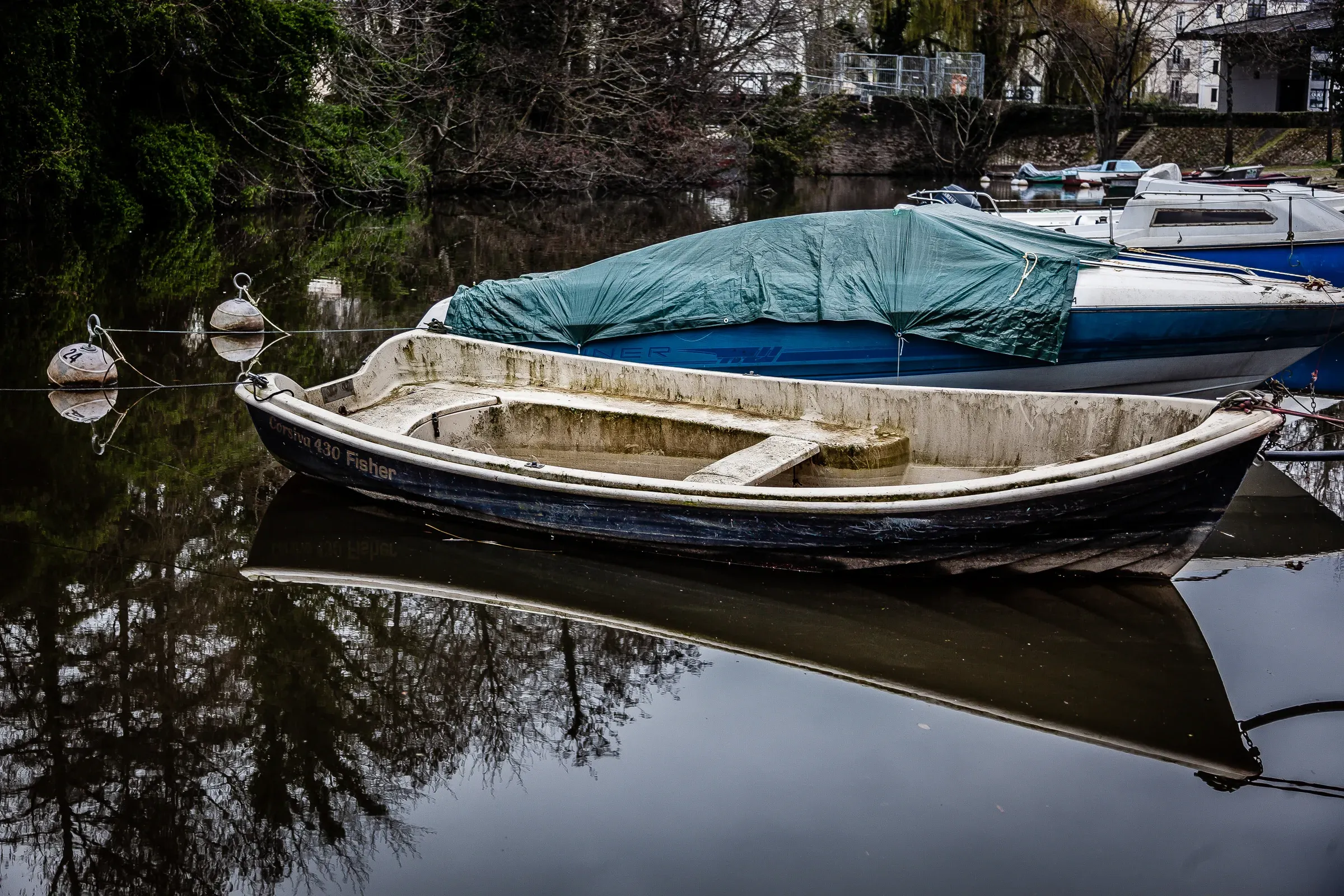 A weathered rowboat with a green tarp, moored on a still dark river with bare winter trees reflected behind, Nantes
