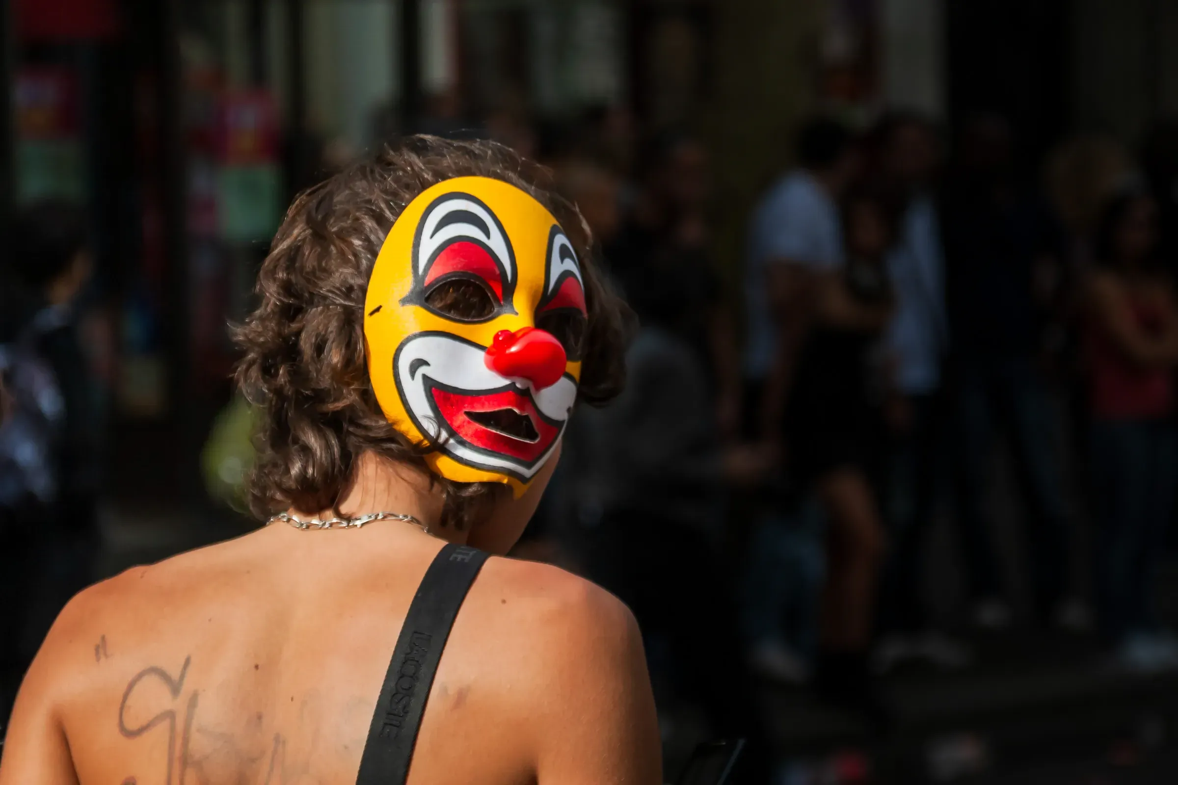 Shirtless person in a yellow clown mask, seen from behind, walking through a sunlit street crowd